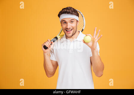 Cheerful young man holding ball et tennis racket sur fond jaune Banque D'Images