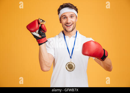 Succès joyeux jeune homme boxer dans les gants rouge et la médaille holding trophy cup sur fond jaune Banque D'Images