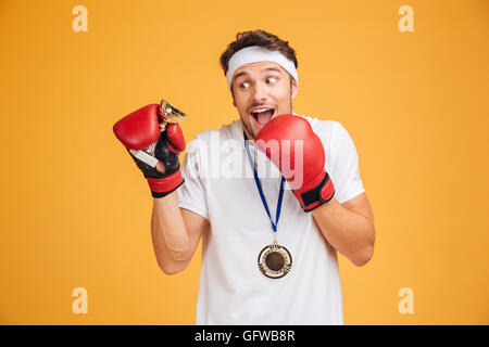 Heureux heureux jeune homme boxer dans les gants rouges avec la coupe et la médaille trophée sur fond jaune Banque D'Images