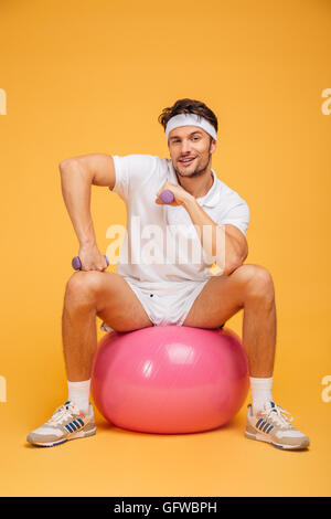 Handsome smiling sportsman assis sur le fitness ball holding dumbbells isolé sur fond orange Banque D'Images