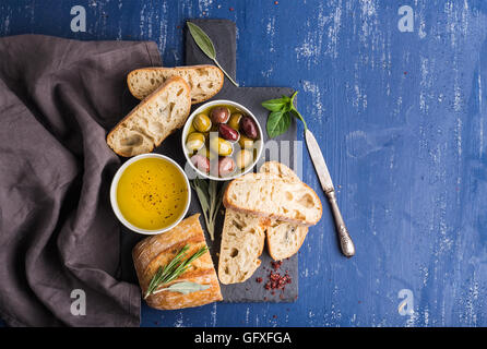 En-cas méditerranéens. Olives, huile, d'herbes et de tranches de pain ciabatta sur ardoise noir peint sur fond bleu foncé Banque D'Images