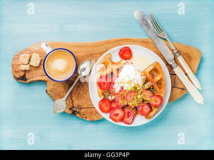 Le petit-déjeuner ensemble. Maison chaleureuse belgique gaufres à la crème fouettée, fraise, sirop d'érable et pistaches concassées, tasse de café, sucre brun olive sur planche de bois rustique sur fond peint bleu Banque D'Images