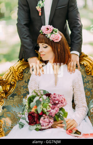 Portrait de rousse charmante épouse modèle avec bouquet de mariage à la main assis sur une chaise, groom de toucher ses épaules par derrière Banque D'Images