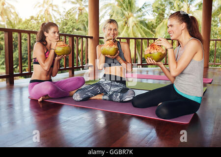 Trois jeunes femmes assis ensemble au cours de yoga et de boire le jus de noix de coco. Groupe de personnes qui prend une pause de la séance d'entraînement yoga. Banque D'Images
