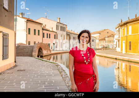 Portrait de l'environnement méditerranéen de mature woman in red dress balade au bord de l'eau canaux d'un vieux village en Italie, Comacchio, connu sous le nom de la Petite Venise Banque D'Images