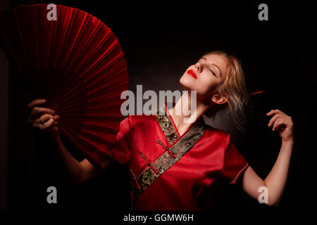 Fille en costume de geisha avec ventilateur rouge sur fond noir Banque D'Images