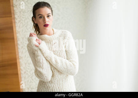 Jolie jeune femme en pull par la fenêtre dans la chambre Banque D'Images