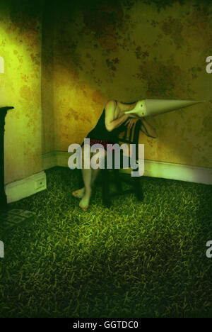 Surreal portrait of young woman wearing hat cône assis sur une chaise dans la pièce remplie d'herbe Banque D'Images