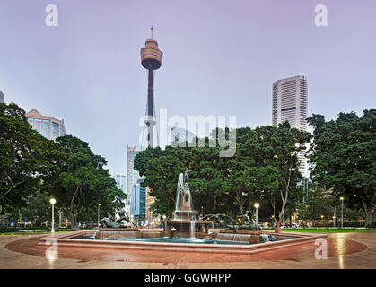 Hyde Park à Sydney au lever du soleil. Fontaine entourée par de vieux grands arbres et de hautes tours en arrière-plan. Banque D'Images