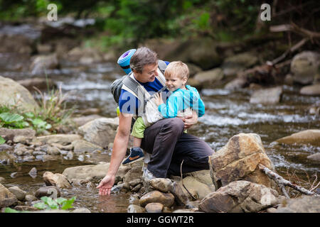 Man de toucher l'eau dans la rivière de montagne tout en portant son fils en wrap sling Banque D'Images