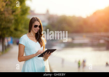 Belle jeune femme brune à l'écoute de la musique dans les écouteurs, holding tablet et profiter de la musique. Vieille ville avec rivière sur b Banque D'Images