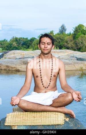 Un jeune prêtre hindou (brahmane), est assis en méditation sur une rive du fleuve. Banque D'Images