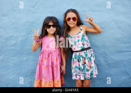 Portrait de deux petites filles se tenant ensemble le port de lunettes gesturing signe de paix contre mur bleu. Filles préadolescentes élégant Banque D'Images