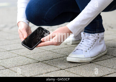 Plan rapproché sur personne agenouillée en jeans et chaussures décrochant broken téléphone sur trottoir pavé en pierre en plein air Banque D'Images