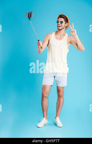 Cheerful young man in hat et lunettes en tenant avec monopode et smartphone selfies Banque D'Images