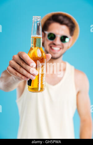 Smiling young man in hat et lunettes vous donnant une bouteille de bière sur fond bleu Banque D'Images