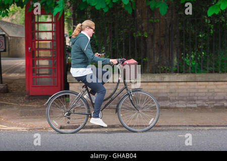 Femme cyclisme, un étudiant regarde son téléphone tout en vélo le long de Trumpington Street dans le centre de Cambridge, Angleterre, Royaume-Uni. Banque D'Images