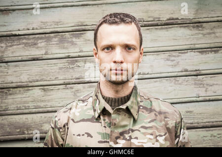 Jeune homme sérieux militaire caucasienne des uniformes de camouflage. Portrait frontal Gros plan sur green wooden wall Banque D'Images