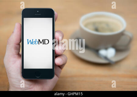 Un homme se penche sur son iPhone qui affiche le logo WebMD, en restant assis avec une tasse de café (usage éditorial uniquement). Banque D'Images