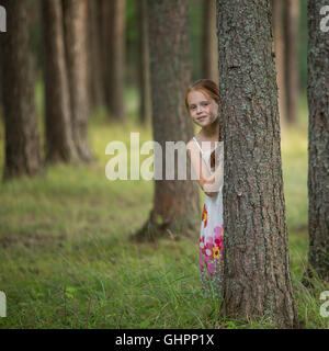 Cute little girl peeking de derrière un arbre dans une forêt de pins. Banque D'Images
