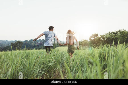 Vue arrière shot of young man and woman holding hands running through grass field. Jeune couple running on meadow sur une journée d'été. Banque D'Images