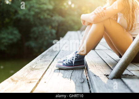 Jeune femme sportive avec de belles jambes assis sur un pont en bois garde-corps en jeans sneakers Banque D'Images