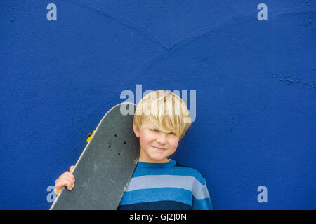 Portrait of cute boy carrying skateboard sur l'épaule en avant du mur bleu Banque D'Images