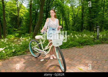 Femme portant une salopette en jean et une blouse ample, debout avec son vélo sur les briques dans un parc Banque D'Images