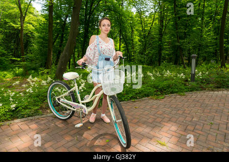 Smiling girl wearing denim bleu de travail et d'un chemisier ample, debout avec son vélo sur les briques dans un parc Banque D'Images