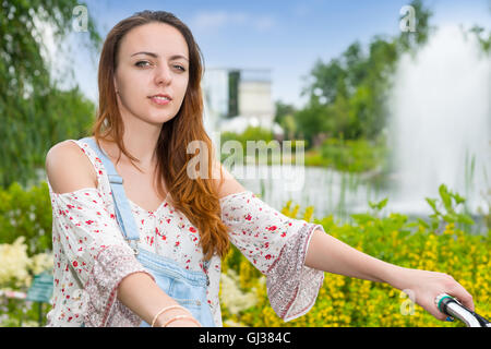 Portrait d'une jeune femme portant une salopette en jean et une blouse ample sur un vélo dans un parc avec fontaine, fleurs colorées, Banque D'Images