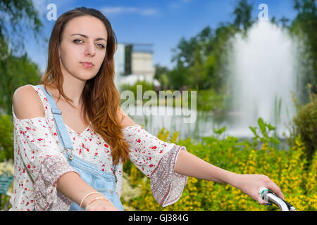 Portrait of a young woman wearing denim bleu de travail et une blouse ample sur un vélo dans un parc avec fontaine, fleurs, buissons et arbres différents dans un arrière-plan. Banque D'Images