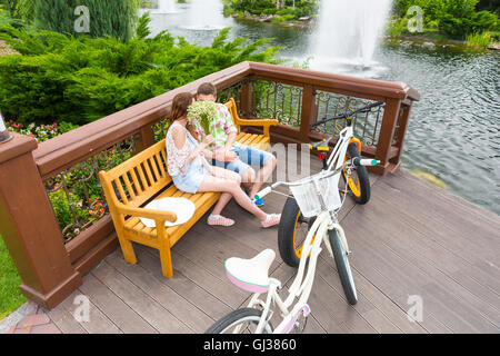 Boy and girl kissing se cacher derrière le petit bouquet de fleurs blanches, assis sur un banc en face de leurs vélos garés dans un parc. Banque D'Images