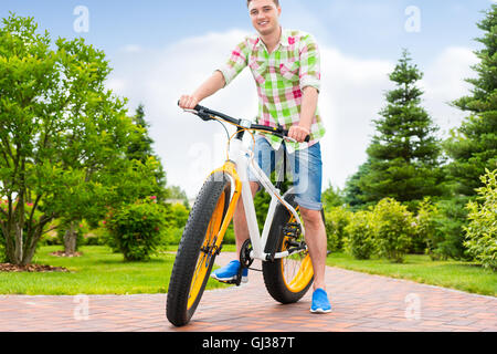 Fabulous bel homme portant une chemise à carreaux verts et rouges assis sur son vélo dans un parc avec de différents arbres d'arrière-plan Banque D'Images