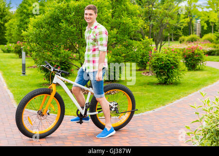 Happy smiling guy portant une chemise à carreaux verts et rouges assis sur son vélo dans un parc avec de différents arbres d'arrière-plan Banque D'Images