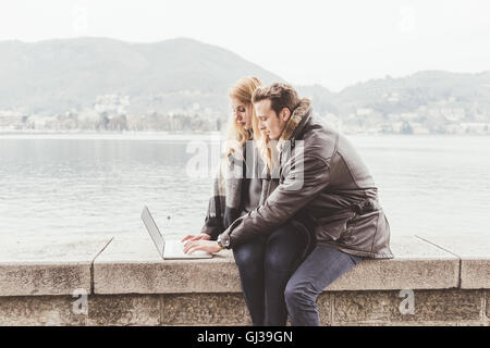 Jeune couple de la saisie sur ordinateur portable sur mur du port, le lac de Côme, Italie Banque D'Images