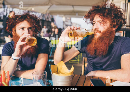 Jeune homme aux cheveux rouges jumeaux hipster et barbes de boire une bière au bar-terrasse Banque D'Images