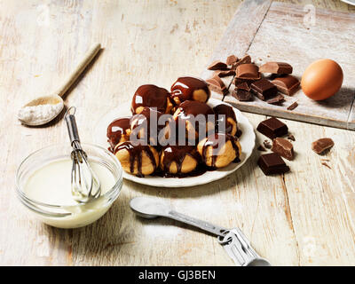 Profiteroles au chocolat chaud, sauce à la crème dans un bol avec un fouet, cuillère en bois avec de la farine, l'oeuf, le sucre en cuillère de mesure Banque D'Images