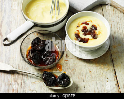 Les pruneaux à la crème anglaise dans un bol blanc, pruneaux à cuillère et bol en verre de sirop, de crème anglaise en pot vintage, table en bois Banque D'Images