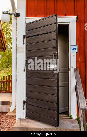 Old weathered et toilettes noir porte en bois à l'angle d'un bâtiment en bois rouge et blanc. Banque D'Images