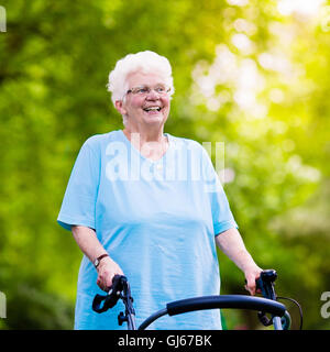 Senior lady handicapés avec un handicap bénéficiant d'une promenade dans un parc ensoleillé poussant sa marchette ou fauteuil Banque D'Images