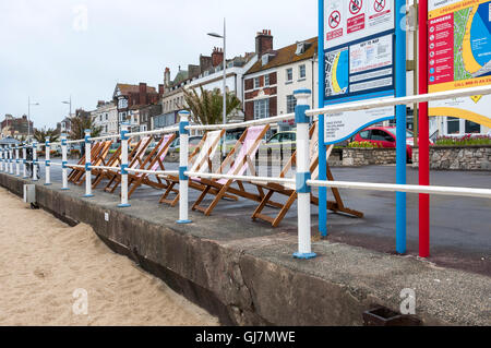 Une rangée de chaises vides partiellement obscurci par des garde-corps bleu et blanc donnent sur une plage de sable fin sur une journée terne à Weymouth Banque D'Images