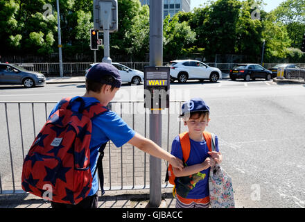 L'école à pied grand frère plus jeune frère aide à attendre signer par passage piétons rue de feux de circulation sur une route très fréquentée en UK KATHY DEWITT Banque D'Images