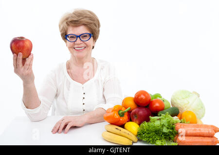 Aîné attrayant Caucasian woman showing red apple, les légumes frais et les fruits sont sur la table, fond blanc Banque D'Images
