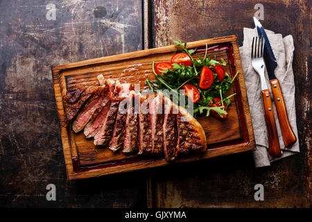 Tranches de boeuf grillé bifteck de contre-filet barbecue et une salade avec des tomates cerises et de la roquette sur une planche à découper sur fond sombre Banque D'Images