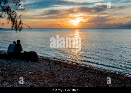 Un couple d'une silhoutte of mountain range at sunset assis sur un journal sous un arbre, profitant du coucher du soleil tropical sur la plage. Banque D'Images