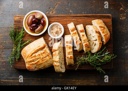 Pain ciabatta frais coupé en tranches sur une planche à découper en bois avec des olives et des herbes sur fond de bois rustique Banque D'Images