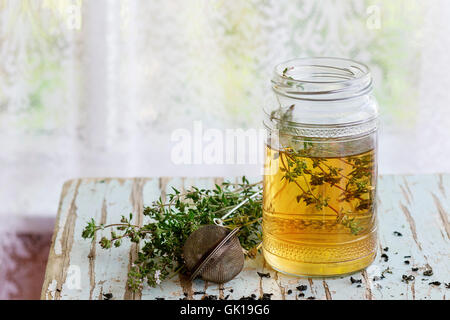 Bocal en verre de tisane chaude avec bouquet de thym frais, servis avec du thé vintage-crépine sur le vieux tabouret en bois avec fenêtre à backg Banque D'Images