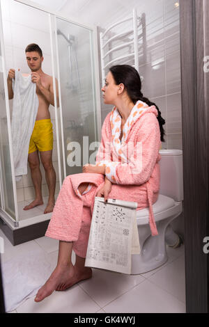 Femme en peignoir assis sur un journal avec toilettes et en attente de son petit ami alors qu'il essuie-tout dans la cabine de douche avec portes en verre transparent dans la salle de bains. Banque D'Images