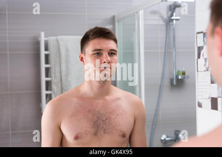 Jeune homme après rasage mauvais debout devant un miroir dans la salle de bains moderne carrelée à la maison Banque D'Images