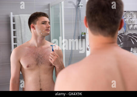 Homme avec des coupures sur son visage après rasage mauvais holding un rasoir alors qu'il se tenait devant le miroir de la salle de bains moderne carrelée à la maison. Banque D'Images